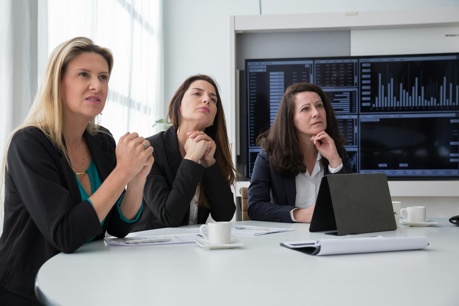 Three women in a business meeting room discussing charts and data on screens.